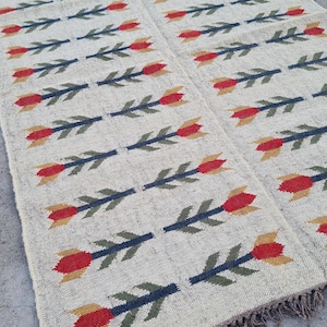 May include: A rectangular beige rug with a repeating pattern of red and yellow tulip-like flowers on dark blue stems. The rug has a fringed edge and is likely made of woven fabric.