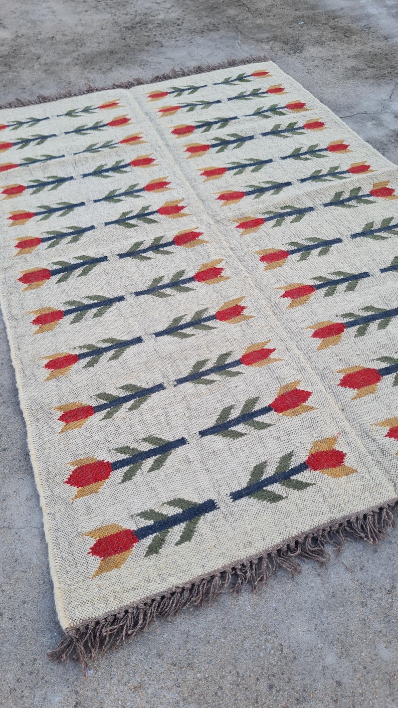 May include: A rectangular beige rug with a repeating pattern of red and yellow tulip-like flowers on dark blue stems. The rug has a fringed edge and is likely made of woven fabric.