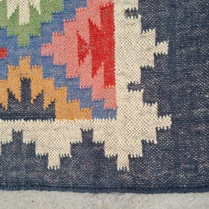 Puede incluir: Una colorida alfombra tejida con un patr&oacute;n geom&eacute;trico. La alfombra presenta un borde azul marino y un dise&ntilde;o central con formas rojas, amarillas, azules, verdes y rosas. La alfombra tiene un borde con flecos.