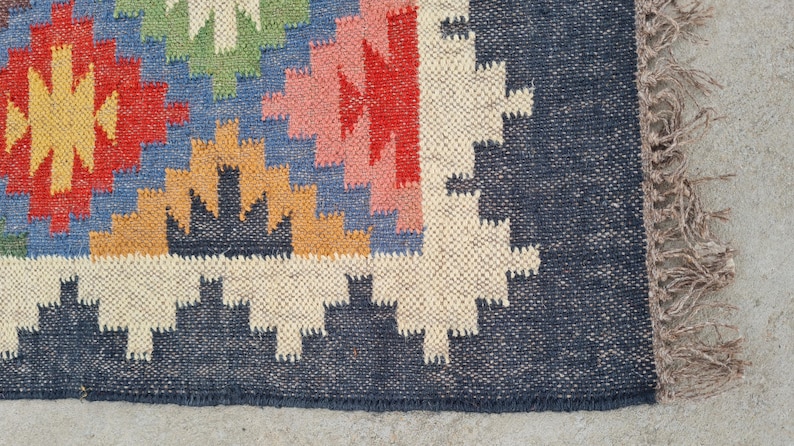 Puede incluir: Una colorida alfombra tejida con un patr&oacute;n geom&eacute;trico. La alfombra presenta un borde azul marino y un dise&ntilde;o central con formas rojas, amarillas, azules, verdes y rosas. La alfombra tiene un borde con flecos.
