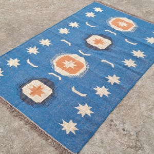 Puede incluir: Una alfombra rectangular azul con un patr&oacute;n repetido de estrellas blancas, lunas crecientes y formas geom&eacute;tricas naranjas y blancas. La alfombra tiene un borde con flecos y est&aacute; colocada sobre una superficie de hormig&oacute;n.