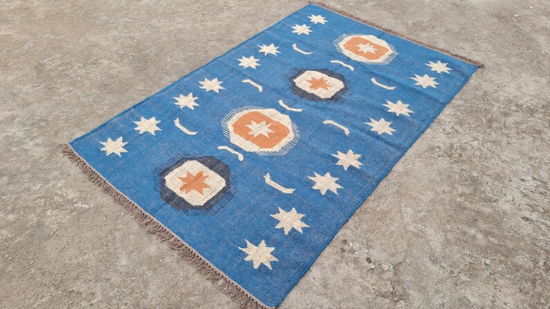 Puede incluir: Una alfombra rectangular azul con un patr&oacute;n repetido de estrellas blancas, lunas crecientes y formas geom&eacute;tricas naranjas y blancas. La alfombra tiene un borde con flecos y est&aacute; colocada sobre una superficie de hormig&oacute;n.