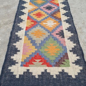 Puede incluir: Una alfombra de pasillo rectangular con un patr&oacute;n geom&eacute;trico en tonos de azul, rojo, amarillo, verde y rosa. La alfombra tiene un borde azul oscuro con un dise&ntilde;o en zigzag blanco y flecos en los extremos. La alfombra est&aacute; enrollada en un extremo.
