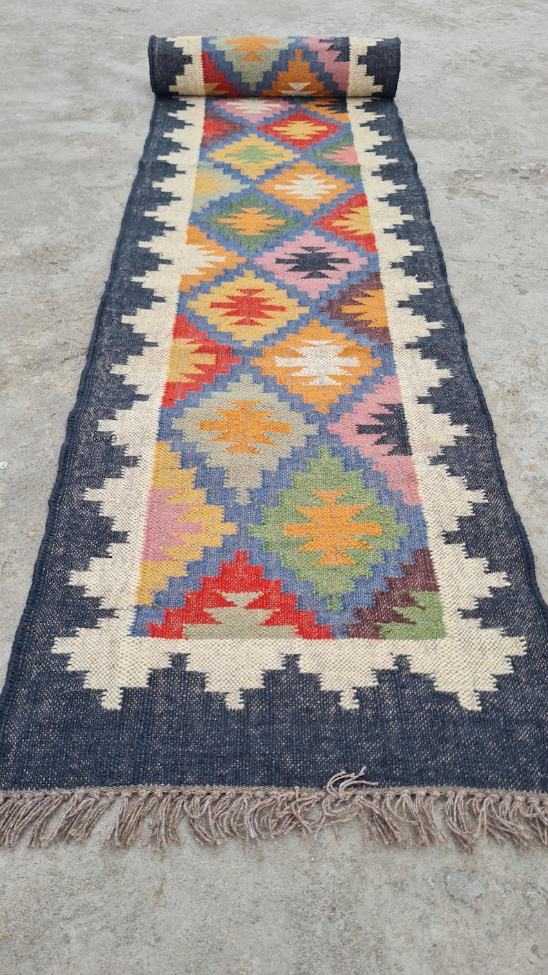 Puede incluir: Una alfombra de pasillo rectangular con un patr&oacute;n geom&eacute;trico en tonos de azul, rojo, amarillo, verde y rosa. La alfombra tiene un borde azul oscuro con un dise&ntilde;o en zigzag blanco y flecos en los extremos. La alfombra est&aacute; enrollada en un extremo.