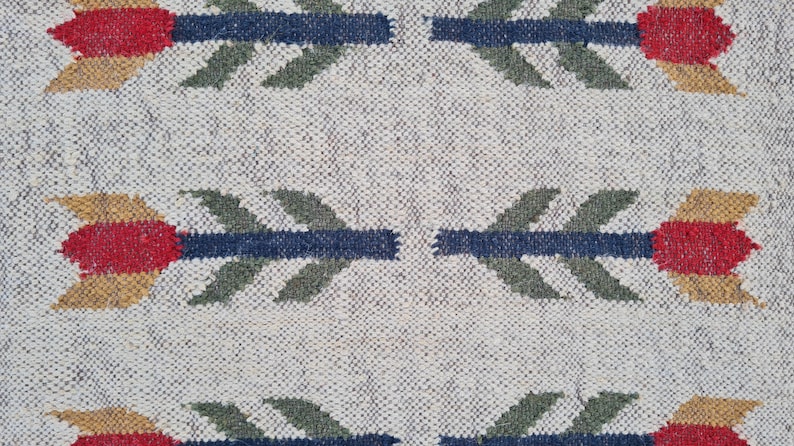 May include: Close-up of a woven textile with a repeating pattern of stylized arrows. The arrows feature red, gold, and blue elements against a neutral, textured background. The textile appears to be made of natural fibers.