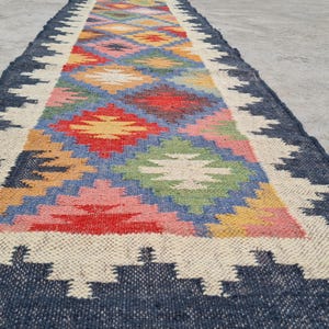 Puede incluir: Una alfombra de pasillo rectangular con un colorido dise&ntilde;o geom&eacute;trico. Tiene un borde azul marino, un borde color crema y un patr&oacute;n central de diamantes en rojo, amarillo, rosa, azul y verde. La alfombra est&aacute; hecha de material tejido.