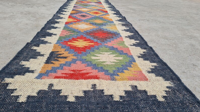 Puede incluir: Una alfombra de pasillo rectangular con un colorido dise&ntilde;o geom&eacute;trico. Tiene un borde azul marino, un borde color crema y un patr&oacute;n central de diamantes en rojo, amarillo, rosa, azul y verde. La alfombra est&aacute; hecha de material tejido.
