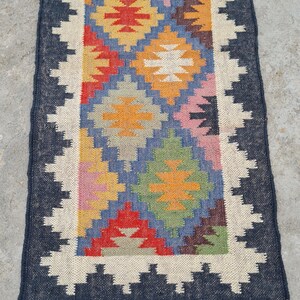 Puede incluir: Una alfombra rectangular tejida a mano con un patr&oacute;n central de formas de diamante coloridas en rojo, amarillo, azul y verde. La alfombra tiene un borde azul oscuro con un borde en zigzag blanco y flecos.