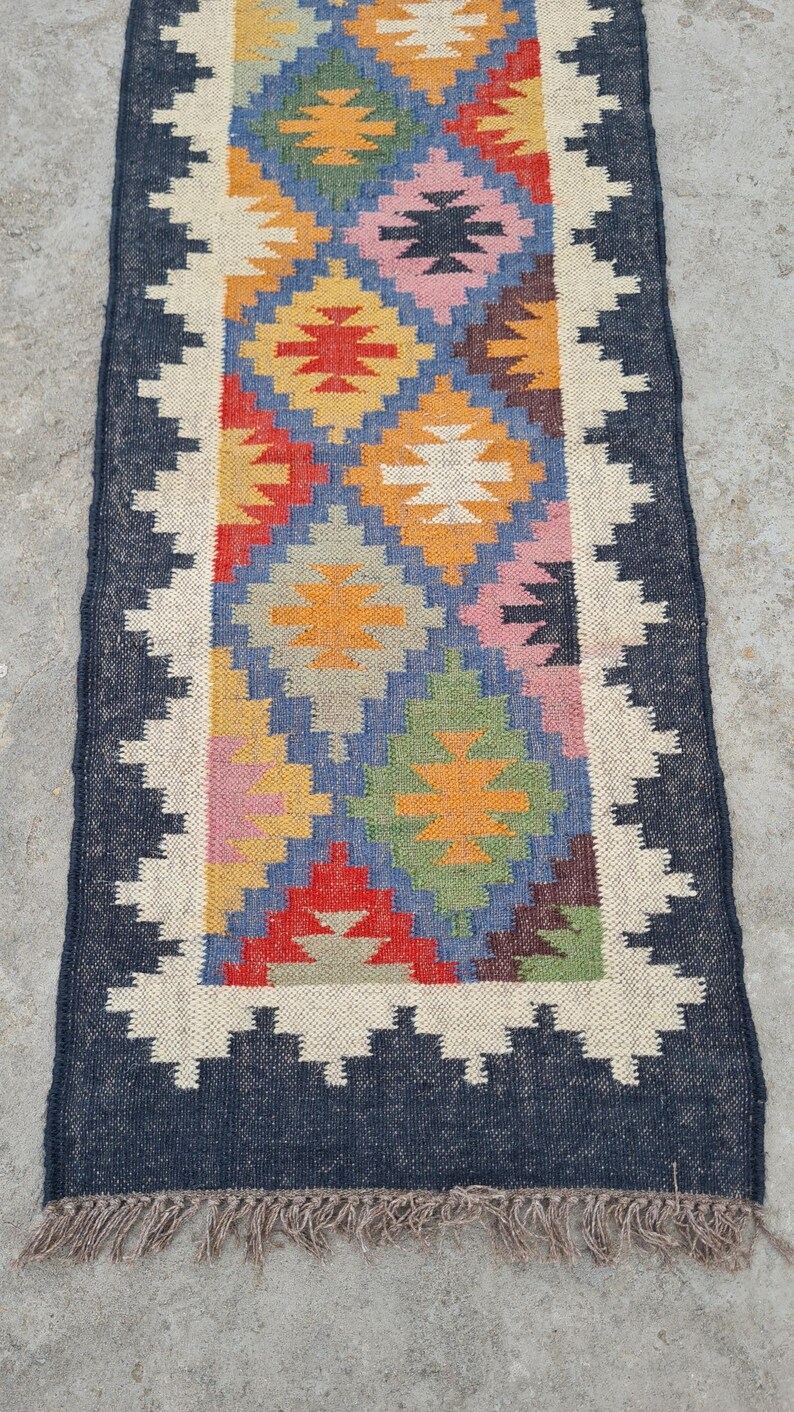 Puede incluir: Una alfombra rectangular tejida a mano con un patr&oacute;n central de formas de diamante coloridas en rojo, amarillo, azul y verde. La alfombra tiene un borde azul oscuro con un borde en zigzag blanco y flecos.