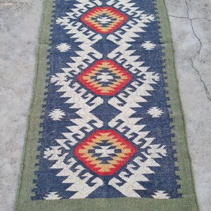 May include: A rectangular runner rug with a geometric pattern. The rug features a navy blue background with white, red, and yellow diamond shapes. The border is green, and the rug has fringe on the ends.