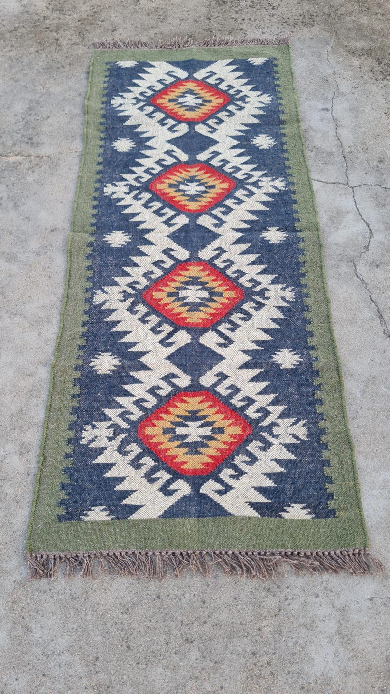 May include: A rectangular runner rug with a geometric pattern. The rug features a navy blue background with white, red, and yellow diamond shapes. The border is green, and the rug has fringe on the ends.