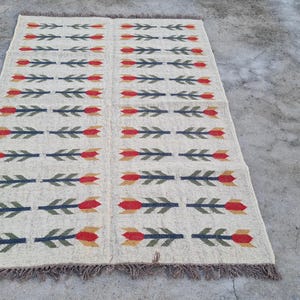 May include: Rectangular beige rug with a repeating pattern of red and yellow tulip-like shapes and green leaves. The rug has a fringed edge and is suitable for a living room or bedroom.