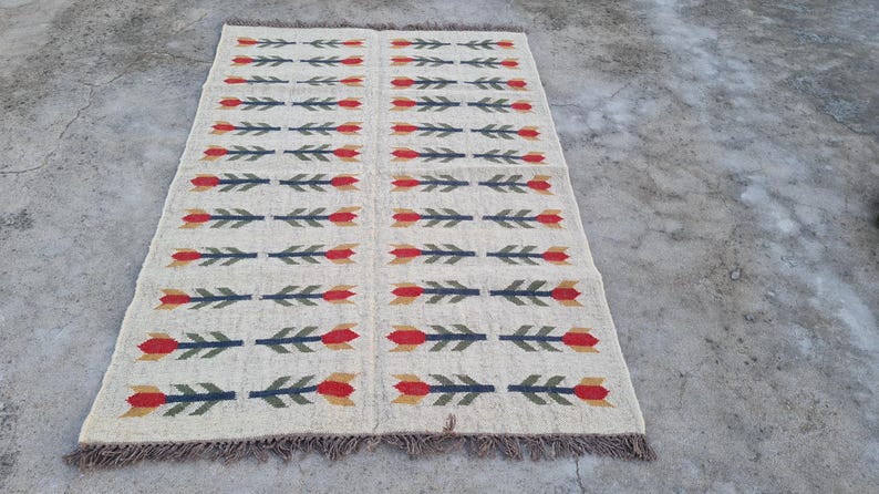 May include: Rectangular beige rug with a repeating pattern of red and yellow tulip-like shapes and green leaves. The rug has a fringed edge and is suitable for a living room or bedroom.