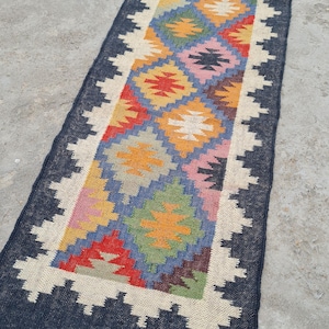 Puede incluir: Una alfombra de pasillo rectangular enrollada con un patr&oacute;n geom&eacute;trico de diamantes multicolores. La alfombra tiene un borde azul oscuro con un borde en zigzag blanco y flecos. El dise&ntilde;o central incluye diamantes rojos, amarillos, verdes y azules.