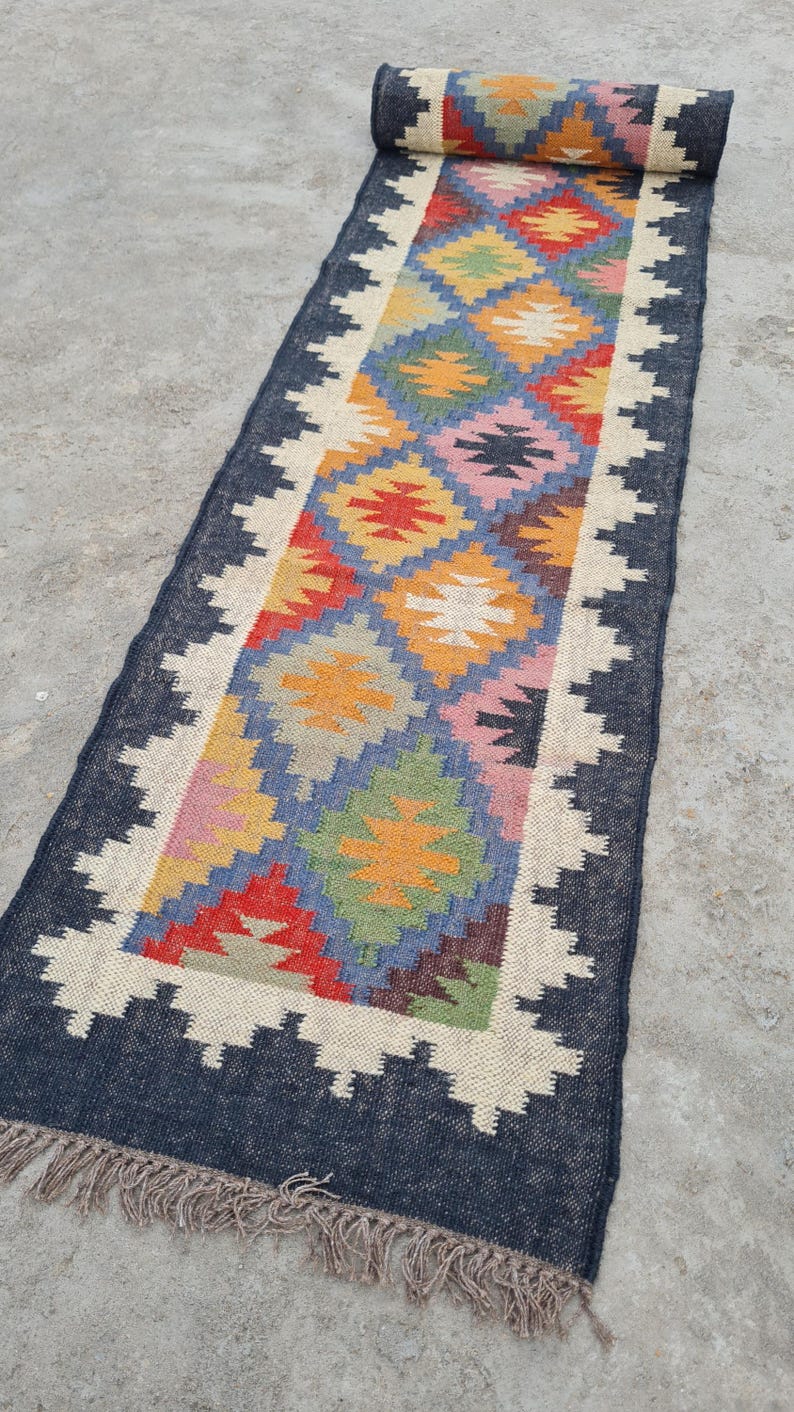 Puede incluir: Una alfombra de pasillo rectangular enrollada con un patr&oacute;n geom&eacute;trico de diamantes multicolores. La alfombra tiene un borde azul oscuro con un borde en zigzag blanco y flecos. El dise&ntilde;o central incluye diamantes rojos, amarillos, verdes y azules.