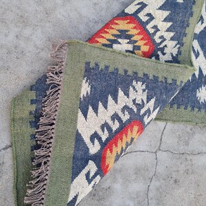 May include: A folded, rectangular rug with a geometric pattern. The rug features a navy blue background with white, red, and yellow geometric shapes. It has a green border and fringed edges.