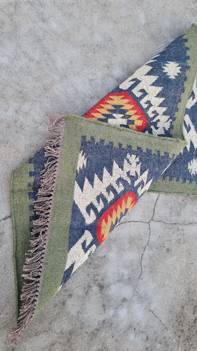 May include: A folded, rectangular rug with a geometric pattern. The rug features a navy blue background with white, red, and yellow geometric shapes. It has a green border and fringed edges.