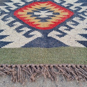 May include: A rectangular rug with a geometric pattern. The rug features a central diamond shape in red and yellow, surrounded by blue and white patterns. The border is green, and the rug has brown fringe.