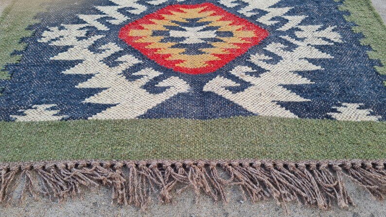 May include: A rectangular rug with a geometric pattern. The rug features a central diamond shape in red and yellow, surrounded by blue and white patterns. The border is green, and the rug has brown fringe.