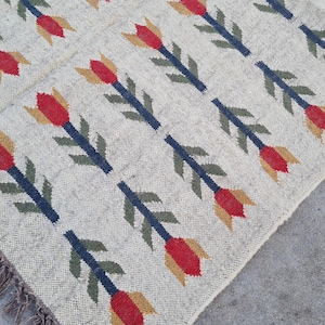 May include: Cream-colored woven rug with a repeating pattern of red, yellow, and green floral motifs. The flowers are arranged on navy blue stems. The rug has a fringed edge.