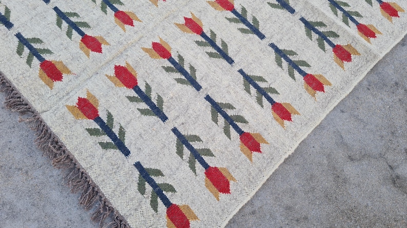May include: Cream-colored woven rug with a repeating pattern of red, yellow, and green floral motifs. The flowers are arranged on navy blue stems. The rug has a fringed edge.