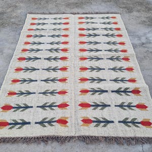 May include: Rectangular beige rug with a repeating pattern of red and yellow tulip-like flowers and green leaves on a dark blue stem. The rug has a fringed edge and is displayed on a concrete surface.