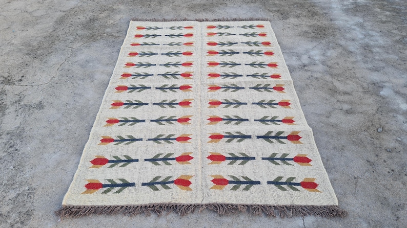 May include: Rectangular beige rug with a repeating pattern of red and yellow tulip-like flowers and green leaves on a dark blue stem. The rug has a fringed edge and is displayed on a concrete surface.