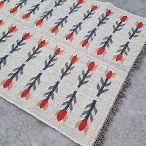 May include: Cream-colored rug with a repeating pattern of red and yellow tulip-like shapes on dark blue stems. The rug has a fringed edge and is made of a woven material.