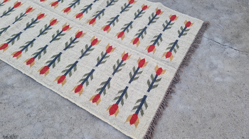 May include: Cream-colored rug with a repeating pattern of red and yellow tulip-like shapes on dark blue stems. The rug has a fringed edge and is made of a woven material.