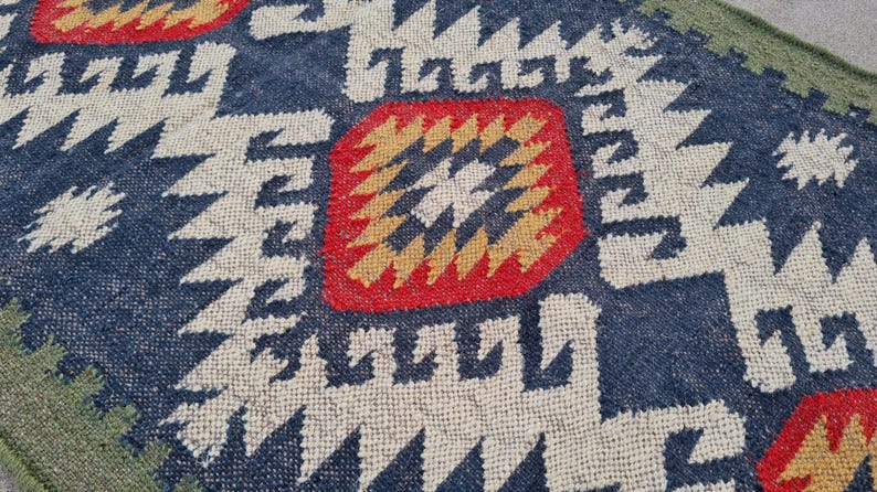May include: Close-up of a woven rug with a geometric pattern. The rug features a navy blue background with white, red, and yellow geometric shapes. The edges are green.