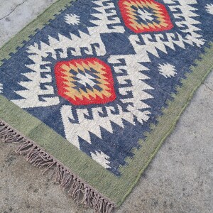 May include: Rectangular blue rug with a geometric pattern in white, red, and yellow. The rug has a green border and fringe on the short ends. The rug is placed on a concrete surface.