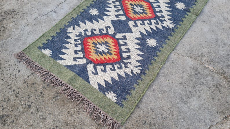 May include: Rectangular blue rug with a geometric pattern in white, red, and yellow. The rug has a green border and fringe on the short ends. The rug is placed on a concrete surface.