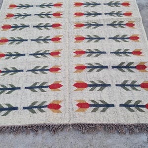 May include: Cream-colored woven rug with a repeating pattern of arrows. Each arrow features a red flower head, green leaves, and a blue shaft. The rug has a fringed edge and is suitable for a living room or bedroom.