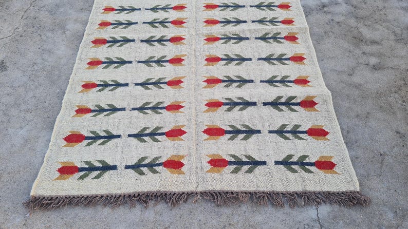 May include: Cream-colored woven rug with a repeating pattern of arrows. Each arrow features a red flower head, green leaves, and a blue shaft. The rug has a fringed edge and is suitable for a living room or bedroom.
