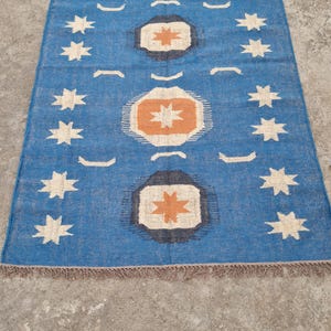 Puede incluir: Alfombra rectangular azul con un patr&oacute;n geom&eacute;trico. La alfombra presenta un dise&ntilde;o repetitivo de estrellas blancas y oct&aacute;gonos naranjas y negros. La alfombra tiene un borde con flecos y est&aacute; colocada sobre una superficie de hormig&oacute;n.