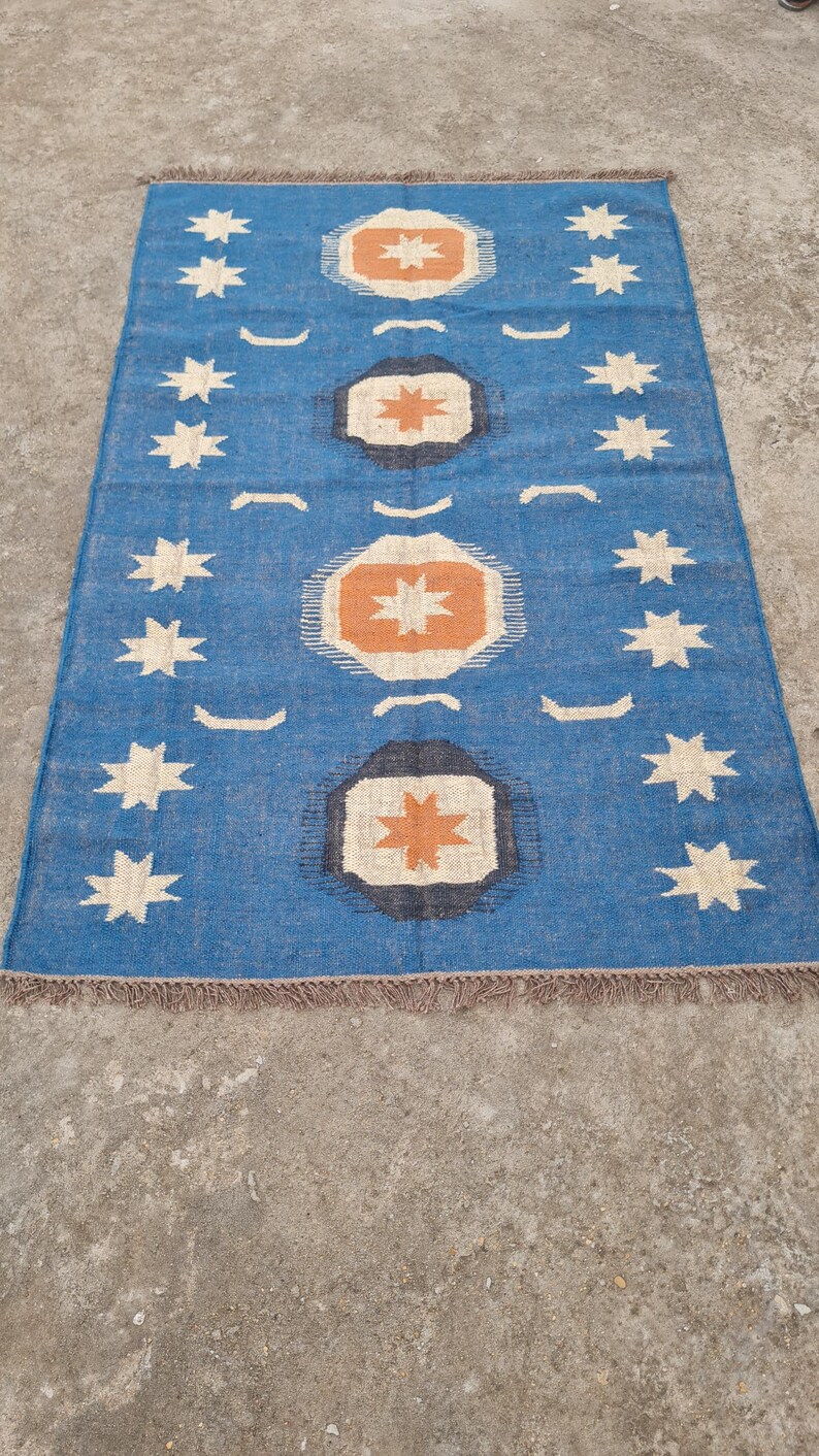 Puede incluir: Alfombra rectangular azul con un patr&oacute;n geom&eacute;trico. La alfombra presenta un dise&ntilde;o repetitivo de estrellas blancas y oct&aacute;gonos naranjas y negros. La alfombra tiene un borde con flecos y est&aacute; colocada sobre una superficie de hormig&oacute;n.