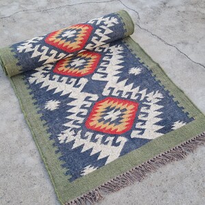 May include: A rolled-up, rectangular rug with a geometric pattern. The rug features a navy blue background with cream and white zigzag designs, and red and gold diamond shapes. It has a green border and fringe.
