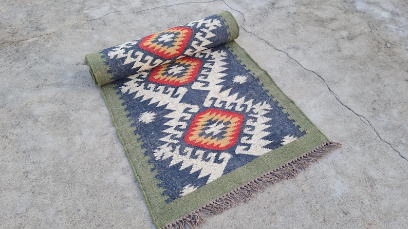 May include: A rolled-up, rectangular rug with a geometric pattern. The rug features a navy blue background with cream and white zigzag designs, and red and gold diamond shapes. It has a green border and fringe.