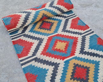 May include: A rolled-up rug with a geometric pattern in red, blue, yellow, and white. The design features repeating diamond shapes with a dark grey border. The rug is made of woven material and has a fringed edge. The rug is suitable for a hallway.