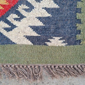 May include: Close-up of a woven rug with a geometric pattern. The rug features a blue, red, and cream design with a green border and brown fringe. The texture of the rug is visible, showcasing the natural fibers.