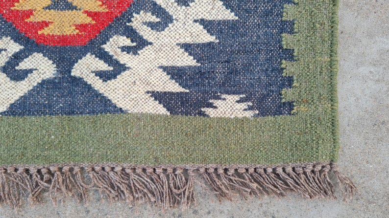 May include: Close-up of a woven rug with a geometric pattern. The rug features a blue, red, and cream design with a green border and brown fringe. The texture of the rug is visible, showcasing the natural fibers.