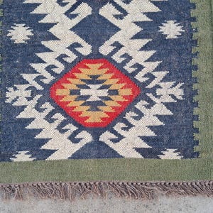 May include: A rectangular rug with a geometric pattern. The rug features a central diamond design in red and gold, surrounded by a navy blue field with white zigzag patterns. A green border frames the design, and the rug has a fringed edge.