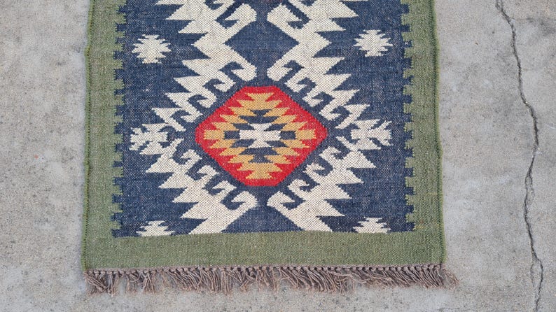 May include: A rectangular rug with a geometric pattern. The rug features a central diamond design in red and gold, surrounded by a navy blue field with white zigzag patterns. A green border frames the design, and the rug has a fringed edge.