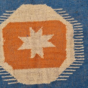 Puede incluir: Alfombra tejida azul con un dise&ntilde;o central que presenta un oct&aacute;gono naranja con una estrella blanca. El oct&aacute;gono est&aacute; rodeado por un borde blanco con una serie de l&iacute;neas cortas y paralelas. El fondo es azul liso.