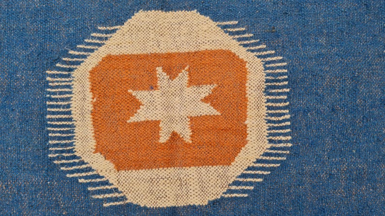Puede incluir: Alfombra tejida azul con un dise&ntilde;o central que presenta un oct&aacute;gono naranja con una estrella blanca. El oct&aacute;gono est&aacute; rodeado por un borde blanco con una serie de l&iacute;neas cortas y paralelas. El fondo es azul liso.
