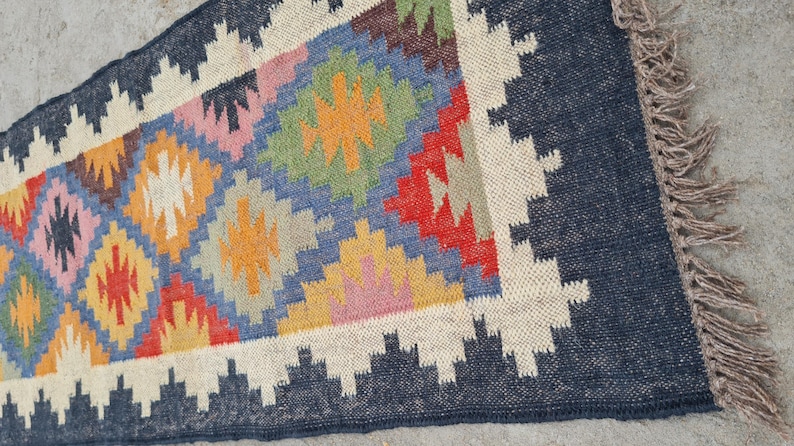 Puede incluir: Una alfombra rectangular con un patr&oacute;n geom&eacute;trico. La alfombra presenta un borde azul marino y un dise&ntilde;o central con diamantes en varios colores, incluyendo azul, naranja, rojo y rosa. La alfombra tiene un borde con flecos.