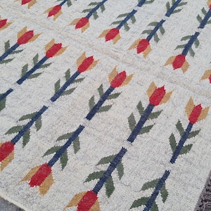 May include: Beige woven rug with a repeating pattern of red, yellow, and blue floral motifs. The rug has a fringed edge and is likely made of natural fibers, suitable for a living room or bedroom.