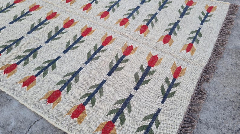 May include: Beige woven rug with a repeating pattern of red, yellow, and blue floral motifs. The rug has a fringed edge and is likely made of natural fibers, suitable for a living room or bedroom.