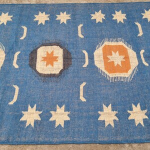 Puede incluir: Una alfombra rectangular azul con un patr&oacute;n repetido de estrellas blancas, medias lunas y formas octogonales con explosiones de estrellas naranjas y beige. La alfombra tiene un borde con flecos.