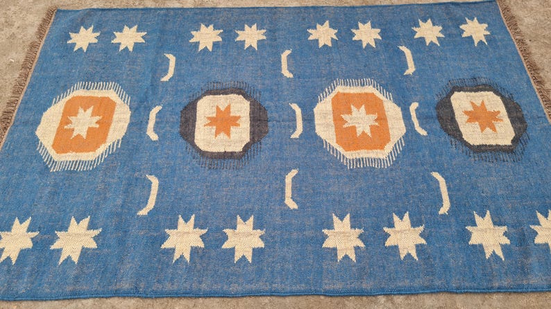 Puede incluir: Una alfombra rectangular azul con un patr&oacute;n repetido de estrellas blancas, medias lunas y formas octogonales con explosiones de estrellas naranjas y beige. La alfombra tiene un borde con flecos.